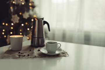 A cup of coffee and a coffee pot with a candlestick stand on the table against the background of a New Year tree