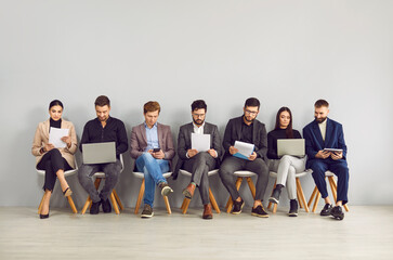 Happy young professional business people, group of applicants are sitting on chairs, waiting for interview, sitting in line with laptops, notebooks and other resources. Staffing employment concept.