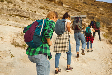Back view of a group of young people tourists with large backpacks hiking in nature climbing a...