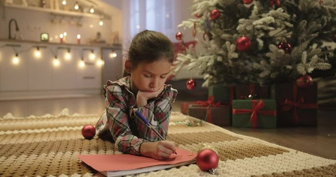 Cute girl writing Christmas letter to Santa Claus lying near decorated Christmas tree