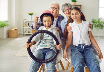 Funny playful grandparents with kids keeping steering wheel sitting on a chairs at home. Child...