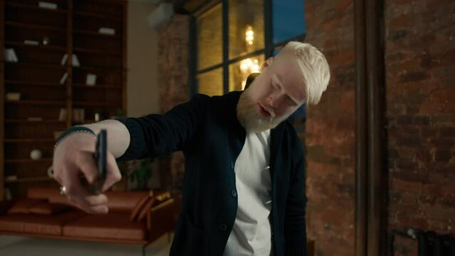 Portrait of handsome albino guy social media blogger making selfie on smartphone at night window. Casual male influencer smiling posing to camera. Trendy man using mobile phone selfie photos in loft