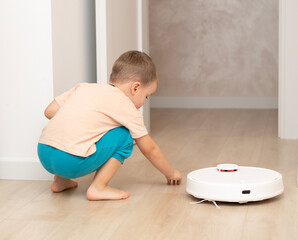 Cleaning concept. A little cheerful boy in home clothes, 4 years old, throws torn paper into small pieces on the floor for cleaning and suction by a white robot vacuum cleaner.