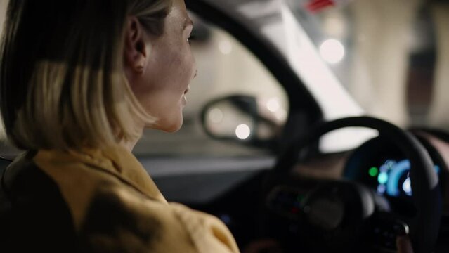 Eco friendly transportation and car insurance. Side view of focused woman driving electric vehicle at night with city lights on background, car interior visible. Attentive auto driver