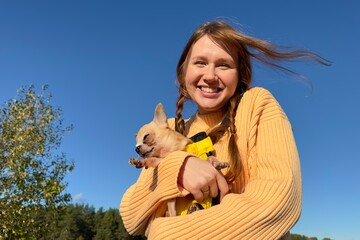 Happy young woman or teenager girl walking with her puppy Chihuahua dog in the forest or park at sunny day, hold dog in dog carrier, kangaroo. Dogs transportation 