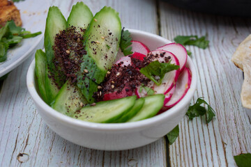 Sliced cucumbers, radishes with sumac spice in white bowl, wooden table