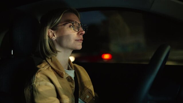 Focused woman in eyeglasses carefully driving at night, illuminated by dashboard lights, concentration, night-time commute on rental electric vehicle. Eco friendly transportation,  car insurance