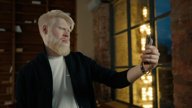 Focused albino entrepreneur talking with coworker during video call in loft office. Surprised businessman discussing project, remote workplace. Freelancer at window with street sign illuminated night