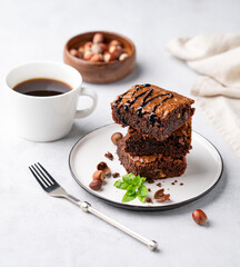 A stack of chocolate brownies with mint, hazelnuts and salted caramel on a plate on a light background with a cup of coffee and nuts.
