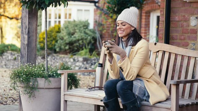 Woman dressed for autumn or winter walk outside house sitting on bench with flask of hot drink - shot in slow motion