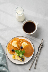 Delicious banana pancakes with fresh fruit slices, berries, mint and honey on a light background with a cup of black coffee, soy milk and shadow.