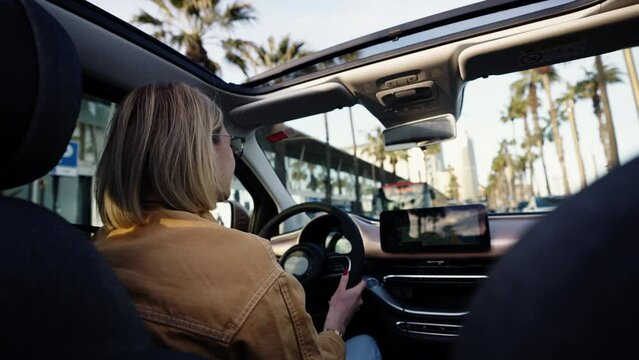 Blonde woman behind the wheel, driving car with sunroof open, palm trees lining the street, concept of urban exploration and modern commuting