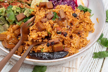 Mexican vegan bowl consisting of red ancho rice, avocado, red cabbage and seitan, greens, radishes. White bowl, wooden table.