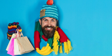 Obraz premium Smiling bearded man in winter clothes with shopping bags looking through paper hole. Handsome male in colourful hat, scarf and gloves with small gift bags. Gifts and presents. Season sales. Discount.