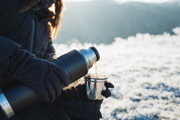 A girl pours hot coffee from a thermos into a mug while sitting in nature in winter, a hot drink...