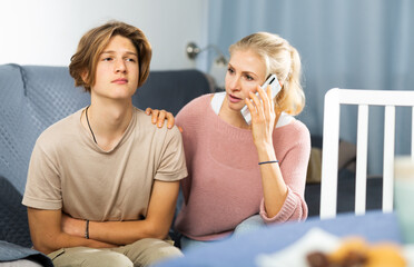 Woman sitting near her sad son who complains of abdominal pain, and calling by phone to doctor