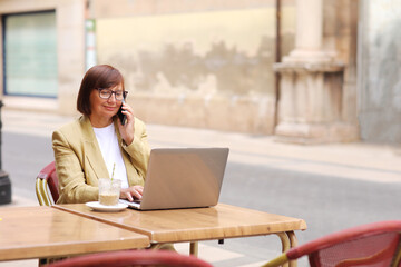 Mature office employee woman talks on smartphone with smile about upcoming business trip while sitting on terrace in cafe with cup of coffee and laptop, happy businesswoman working remotely online.