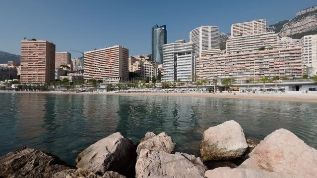The famous beach Larvotto of principality of Monaco in sunny day, luxury residential complex, sun umbrellas, palm trees, azure water