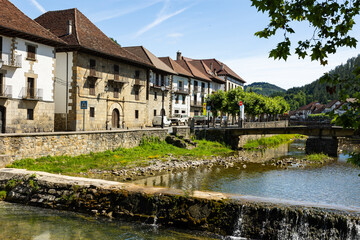 Fototapeta na wymiar Ochagavia townscape on sunny summer day overlooking typical residential buildings with steep brown tiled roofs along stone embankment and bridge across Anduna river on sunny day, Navarra, Spain