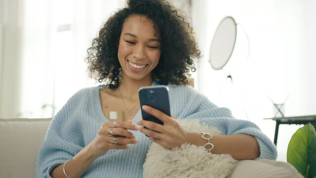 Happy diverse female shopper using instant easy mobile payments, customer making purchase online banking app. Smiling multi racial woman holding smartphone and credit card sitting on couch modern home