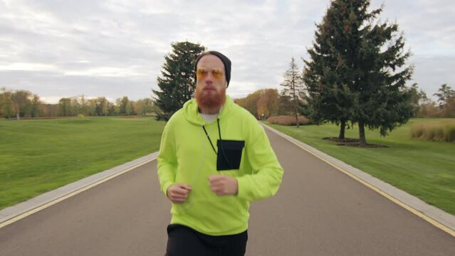 Young male wearing bini hat, tracksuit, in yellow sun glasses, jogging on a fine autumn day in a park with green lawns, multicolored trees, conifers. High quality 4k footage