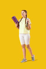 Sporty young woman with skipping rope and fitness mat on yellow background