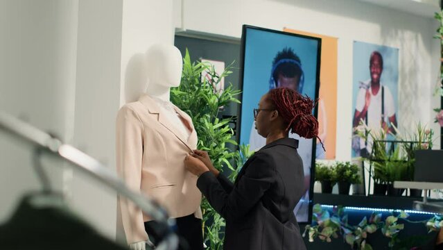 Store assistant looking at formal suit on mannequin in clothing store, closing down buttons. African american retail clerk preparing new fashion collection of stylish blazers