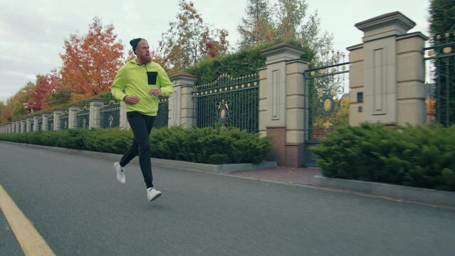Young male runner in sportswear and bini running on asphalt road near exquisite metal fence with multicolored autumn trees, row of evergreen trees behind. High quality 4k footage