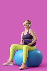 Beautiful young woman with foam roller and headphones sitting on fitness ball against pink background