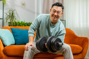 Asian man weight lifting dumbbells, training biceps, workout, burning fat calories, training strength practicing effort domestic fitness aerobics at home room apartment. Sporty adult guy sits on couch