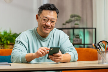 Asian man sits on couch uses mobile phone smiles at home room apartment. Middle-aged Chinese guy texting share messages content on smartphone social media applications online watching relax movie - Powered by Adobe
