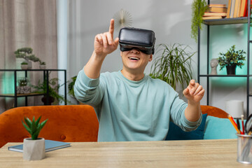 Asian man using virtual reality futuristic technology VR app headset helmet typing on virtual keyboard, play simulation 3D 360 online video game at home apartment. Guy in goggles sitting on sofa