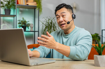 Asian man wearing headset, freelance worker, call center or support service operator helpline, having talk with client or colleague communication support at home room. Guy sits on couch at table desk