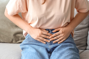 Young woman sitting on sofa and suffering from abdominal pain at home - obrazy, fototapety, plakaty