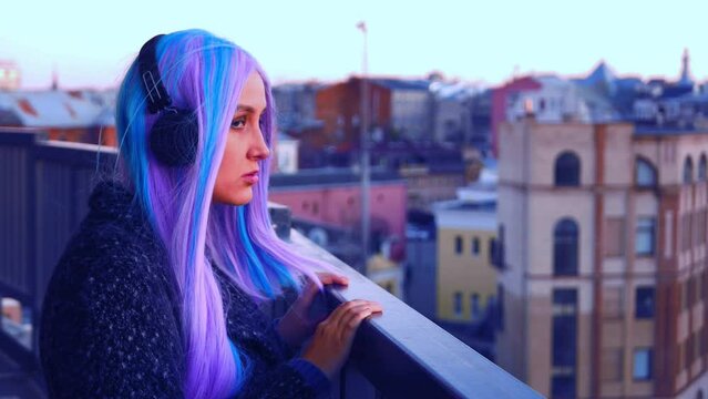 A white girl with long colored hair in dark headphones in a dark coat looks from the balcony at the city 