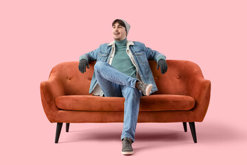 Young man in winter clothes sitting on red sofa against pink background