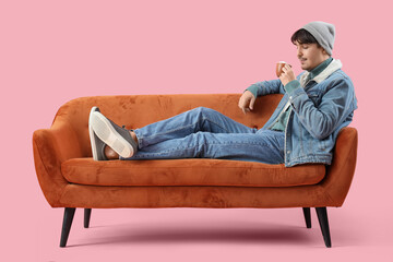 Young man in winter clothes with cup of cocoa lying on red sofa against pink background