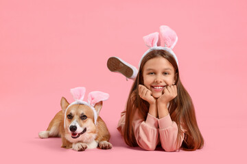 Little girl with cute Corgi dog in bunny ears lying on pink background