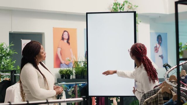Customer helped by employee in SH clothing shop, choosing between shirts using digital screen. Client looking at new collection of garments in discount store using kiosk under retail clerk guidance