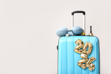 Suitcase with figure 2024 made of foil balloons, travel pillow and Christmas bell near light wall