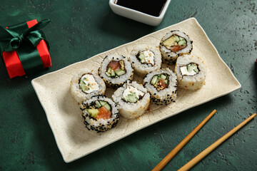 Plate with tasty sushi rolls, soy sauce and gift box for Christmas party on green background, closeup