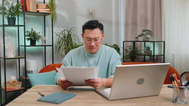 Stressed Asian Man Working On Laptop At Home Room Looking At Unpaid Bank Debt Bills Doing Paperwork Planning Budget Calculate Finances Mortgage Payments. Displeased Guy Throws Paper Bills Overhead