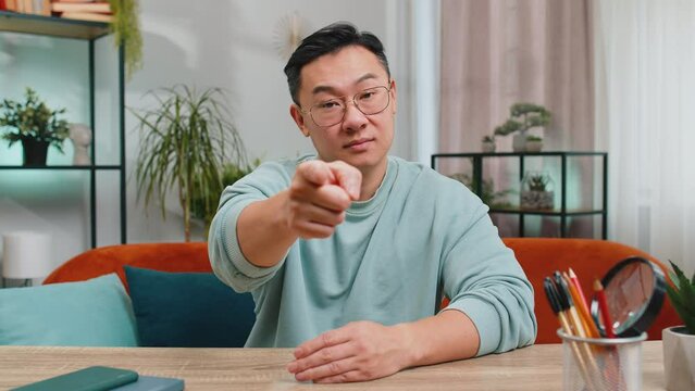 Hey you. Strict unhappy Asian man pointing hand finger to camera, looking angry, making choice, choosing guilty, noticing suspicious, accusing for mistake at home room apartment. Guy sits on couch