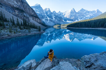 Female tourist carries a backpack, travels, walks, climbs, sits and admires the beauty of the reflection in the water of moraine Lake, Canadian Rocky Mountains - obrazy, fototapety, plakaty