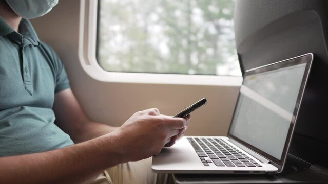 Man texting on phone holding laptop, commuter train in protective mask during coronavirus pandemic. Theme travel by rail, new rules life during COVID 19. - Powered by Adobe