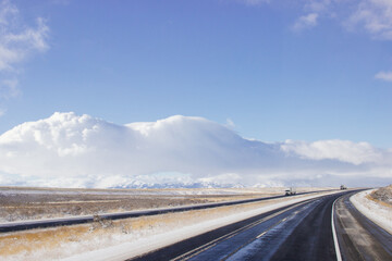 Fototapeta premium Steep highway among snow-capped mountains on a sunny winter day. Bright blue winter sky with large clouds over an asphalt road. Highways in USA. American roads