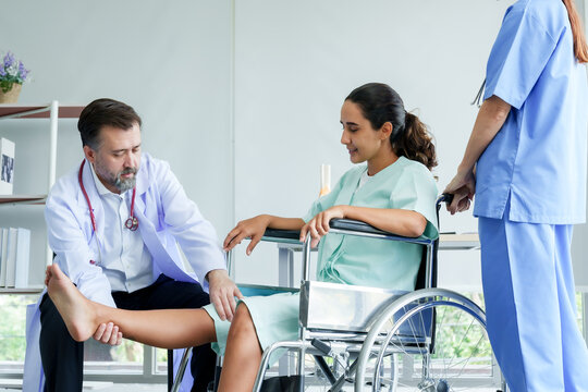 Orthopedic Specialist Lifting Leg Of Female Patient Sitting In A Wheelchair. Check For Injuries After A Broken Leg In The Examination Room. To Prepare For Physical Therapy With A Physical Therapist