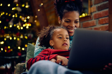 Small black girl and her mother using laptop while relaxing at home on Christmas.