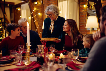 Happy senior woman serving Christmas pie to her family at dining table. - obrazy, fototapety, plakaty