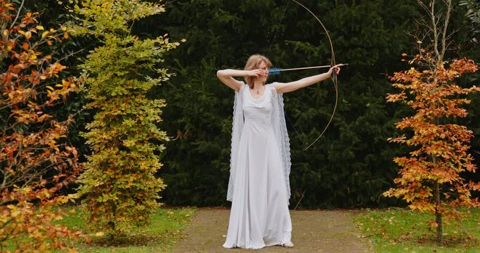 Woman depicting Artemis in a log white dress aiming her bow and looking towards viewer.